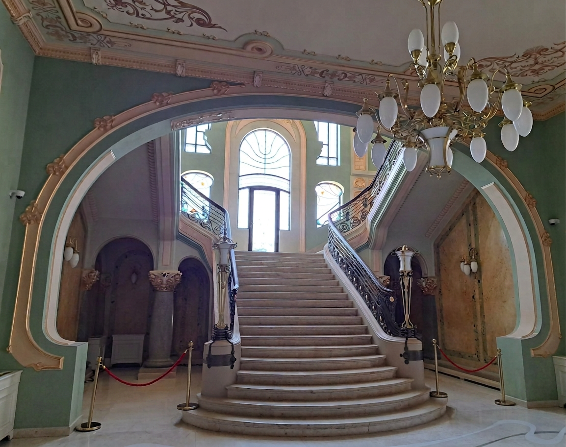 WhatsApp Image 2026-09-08 at 22.06.33 (31) A grand staircase with a decorative railing leads up to large windows, illuminated by a chandelier, within an elegantly styled lobby featuring intricate architectural details and muted green walls.