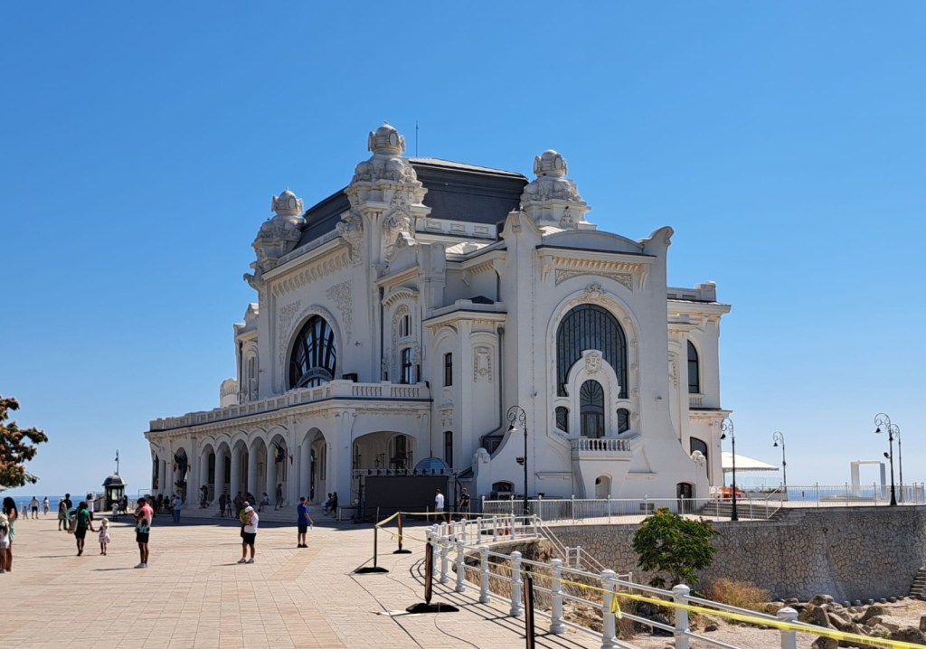 Historic building with ornate architecture, featuring a large arched window and decorative elements, set against a clear blue sky. People walking and enjoying the area in front of the structure.