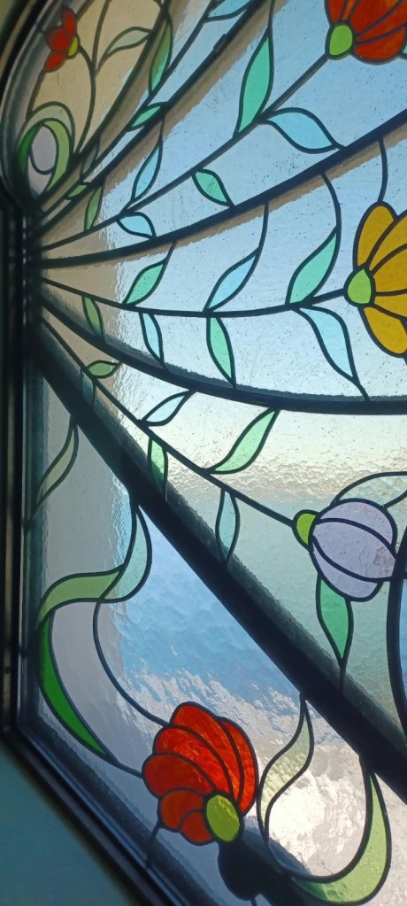 Close-up of a stained glass window featuring colorful floral designs in red, orange, yellow, and purple, with green leaves against a light backdrop.