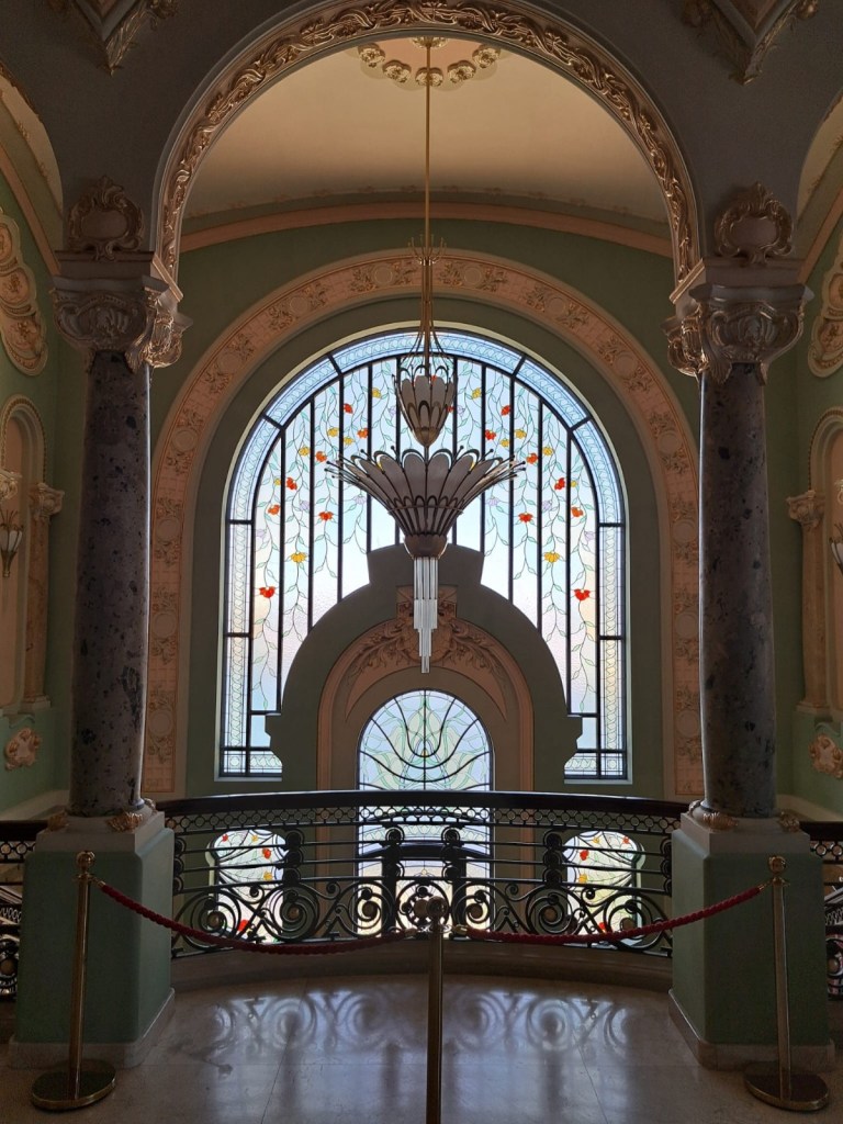 Interior view of a grand staircase featuring ornate architecture, stained glass windows with floral designs, and an elegant chandelier.