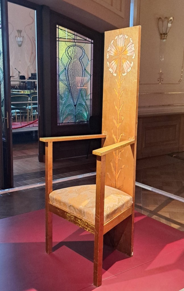A wooden chair with a tall backrest featuring floral designs, set against a backdrop of stained glass windows.