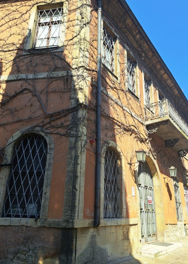 A corner view of an old building with orange walls, featuring a wrought iron balcony and ivy vines climbing the exterior. The building has multiple diamond-pane windows and a heavy door with a security sign.