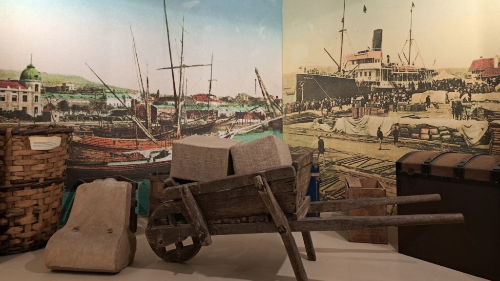 A vintage wooden wheelbarrow placed in front of historical maritime-themed murals featuring ships and a busy port scene.