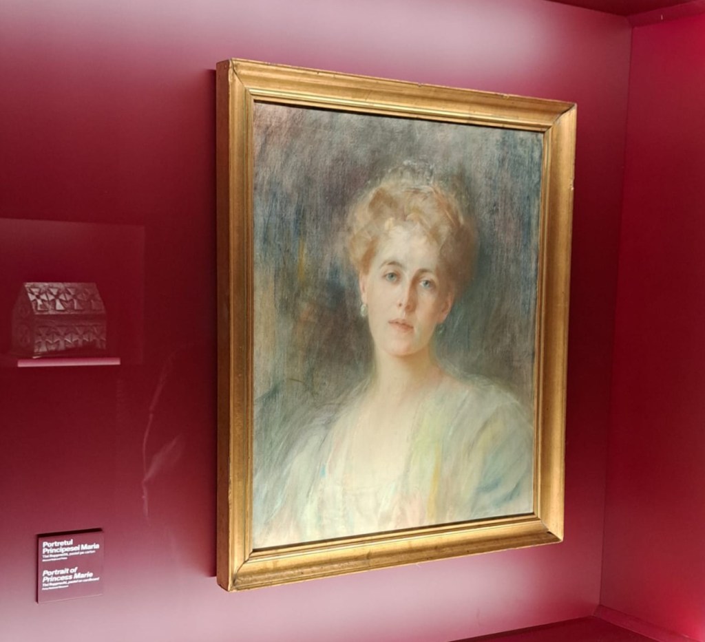 A soft portrait of a woman with light hair and a gentle expression, framed in gold, displayed against a maroon background. A small decorative box is visible in the background.