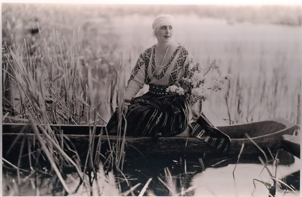 A woman in traditional attire sitting in a canoe, holding flowers, surrounded by tall grasses and a serene water backdrop.