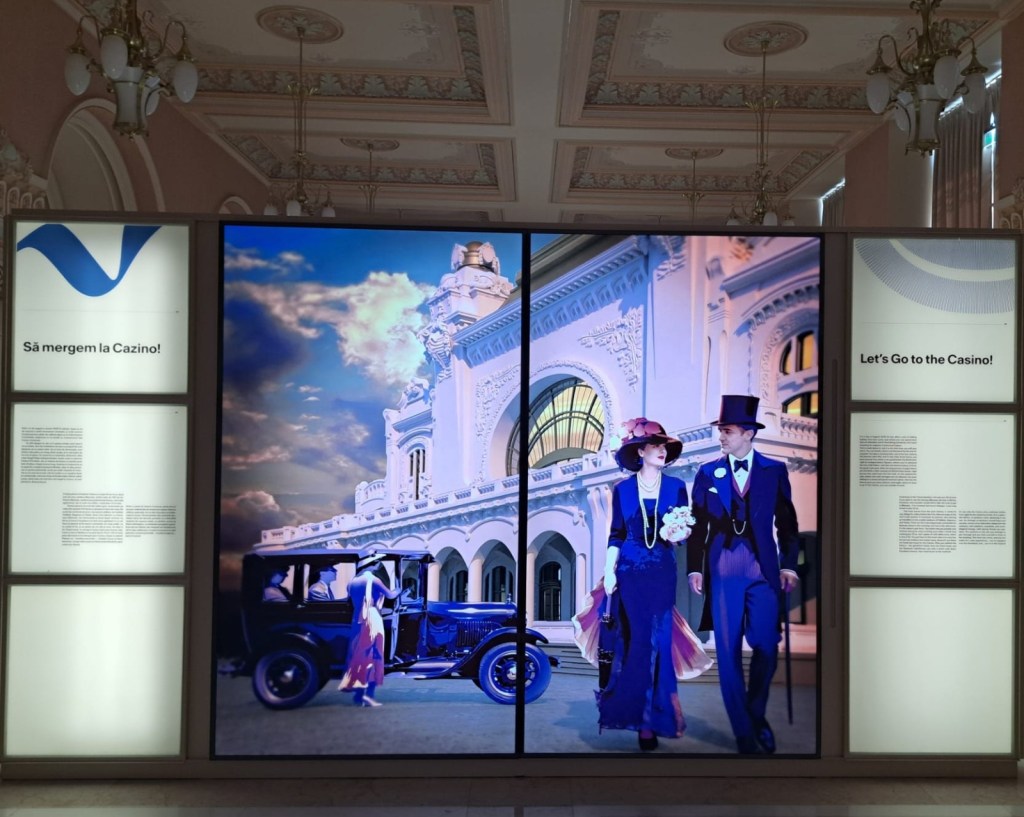 A digital display inside a building featuring a couple dressed in vintage attire walking towards a casino, with an old car parked nearby and an ornate facade in the background. Text on the display includes 'Let's Go to the Casino!' in English and 'Să mergem la Cazino!' in Romanian.
