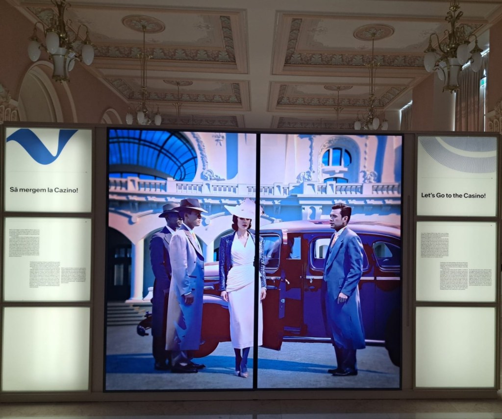 A vintage-themed display featuring four individuals dressed in 1930s attire, standing next to a classic car outside a building with ornate architecture. There are text panels on either side that read 'Să mergem la Cazino!' and 'Let's Go to the Casino!'.