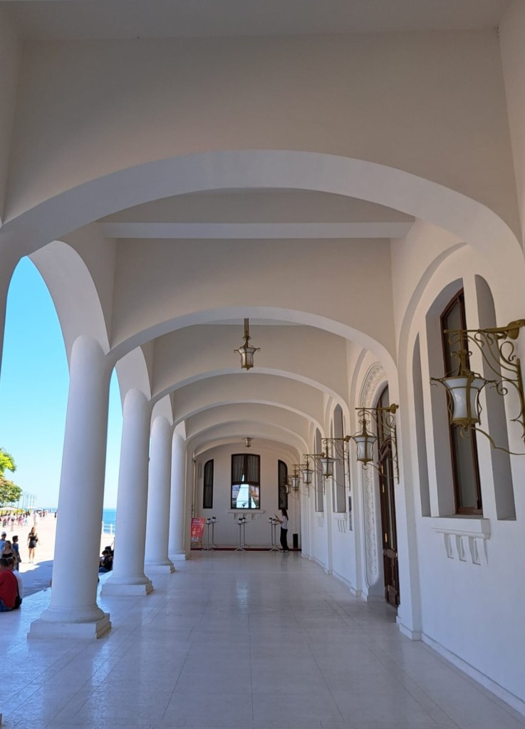 View of a white arched hallway with lanterns, leading toward an entrance, with a person standing near the far end and beach visible in the background.