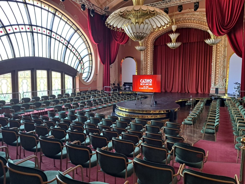 An elegantly styled theater interior with numerous green chairs arranged in rows facing a stage. The stage features a red curtain and decorative elements, with a backdrop displaying "CAZINO MOI AMOUR". Large stained glass windows create an ambient light.