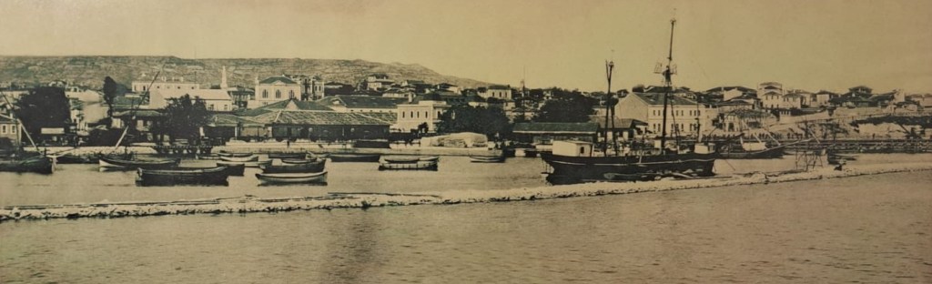 A historic black-and-white photograph of a waterfront scene featuring several boats docked along the shore, with a town visible in the background and a hillside beyond.