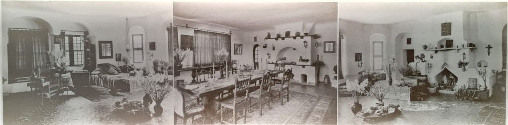 A vintage photograph showcasing a spacious living room with a cozy seating area, a dining table with chairs, and various decorations including plants and artwork. The room features large windows with curtains and a fireplace, creating a warm and inviting atmosphere.