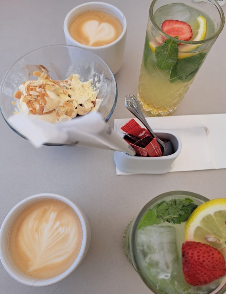 A top-down view of a table with two cups of coffee, one featuring a heart design in the foam, a dessert bowl with cream and almonds, and a glass of refreshing drink with mint, lemon, and strawberry.