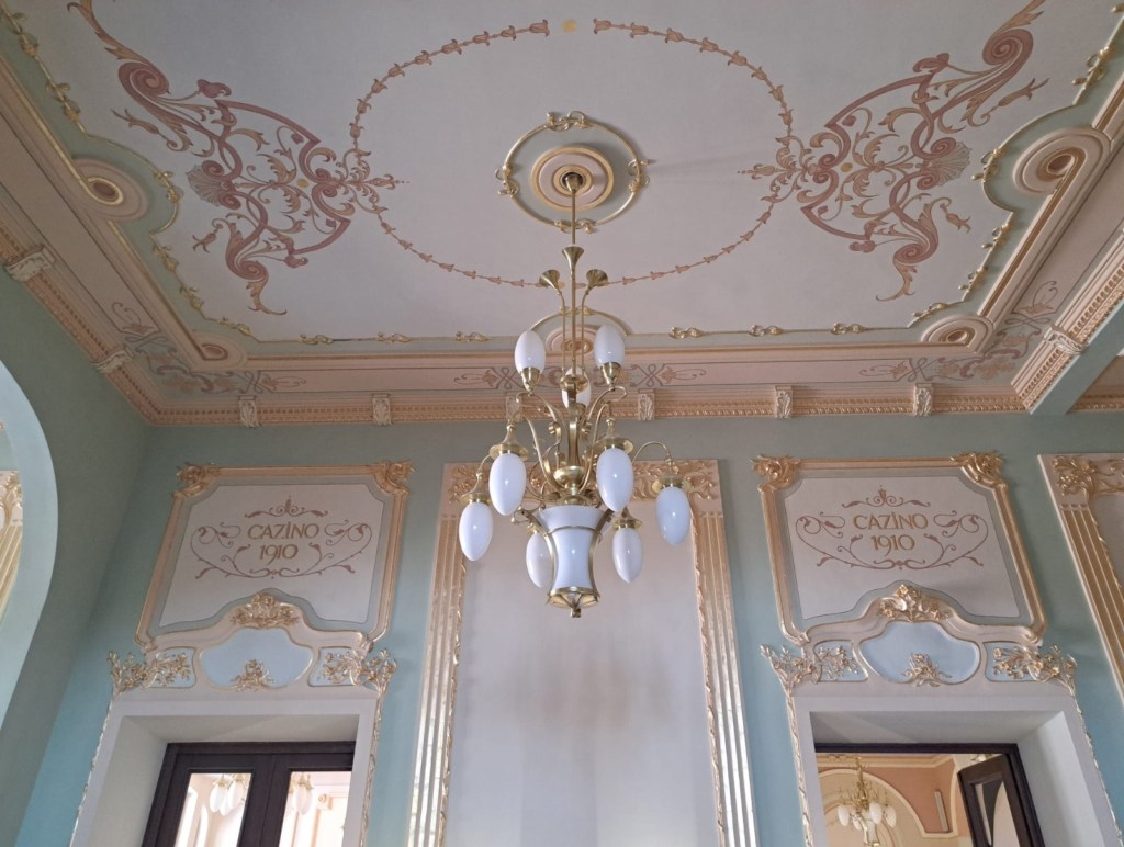 Ornate ceiling with decorative patterns and a chandelier in a historic interior. The walls feature elegant moldings and inscription 'Cazino 1910'.