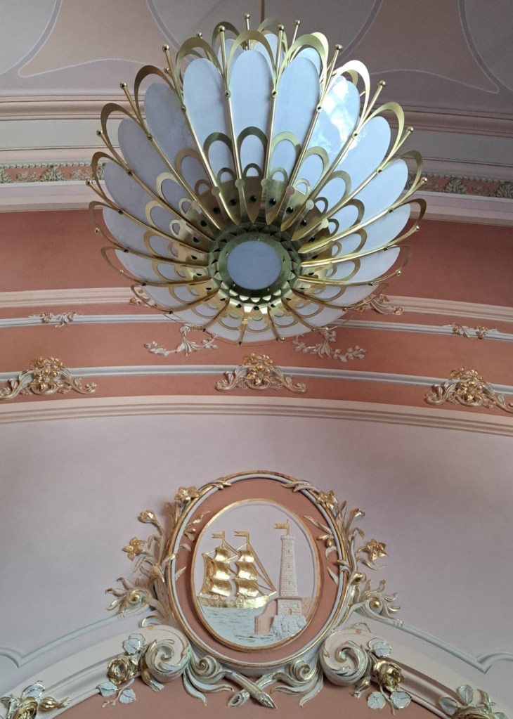 A decorative chandelier resembling a flower, hanging from the ceiling, with a gold and white color scheme, accompanied by ornate wall embellishments featuring a ship and lighthouse.