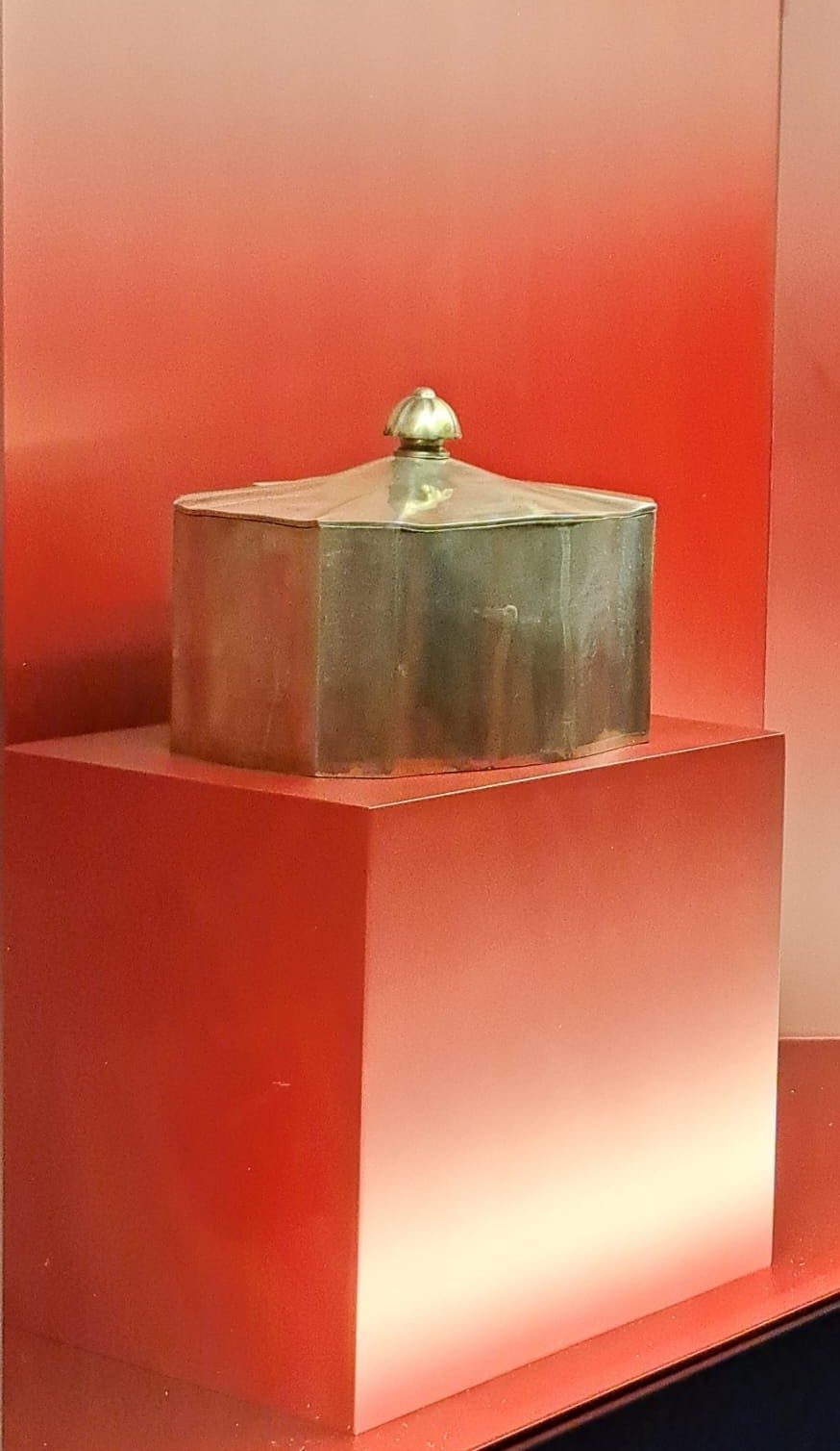 A metallic, octagonal box with a rounded lid displayed on a red pedestal against a gradient red background.