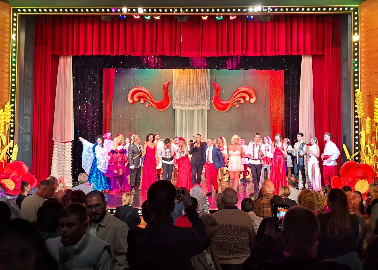 A stage performance featuring a diverse group of actors in colorful costumes, standing together in front of a vibrant backdrop with red curtains and decorative elements. The audience is visible in the foreground, capturing the moment with their phones.