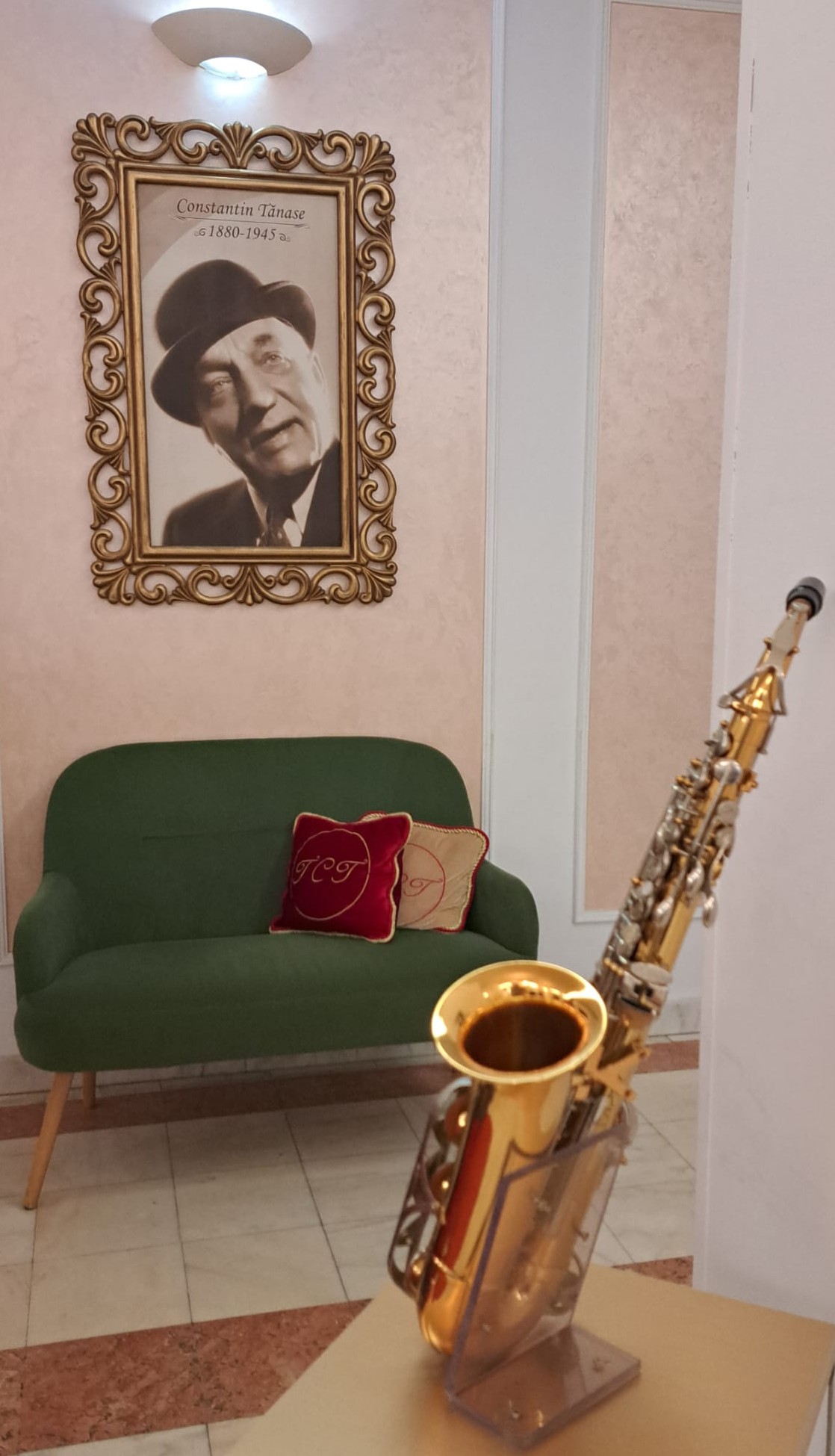 A vintage photograph of Constantin Tânase framed on a pink wall, above a green sofa with red decorative pillows, and a saxophone displayed on a stand.